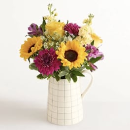 Bouquet of sunflowers, roses, and purple blooms in a white pitcher