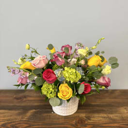 Low arrangement of mixed roses and other blooms in pink, yellow, and red in a white vase