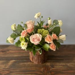Short rustic arrangement of peach roses and white flowers in a bark-covered vase on a wooden table