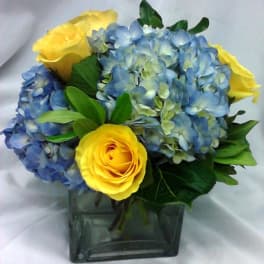 Blue hydrangeas and yellow roses in a square glass vase