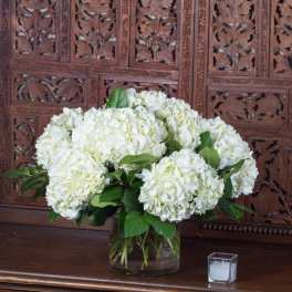 White hydrangea arrangement in a clear glass vase with a small candle beside it
