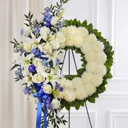 Standing white and blue floral wreath with roses, lilies, and mums on a metal easel
