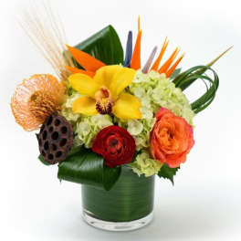 Colorful tropical arrangement with hydrangeas, orchid, rose, and protea in a leaf-wrapped glass vase
