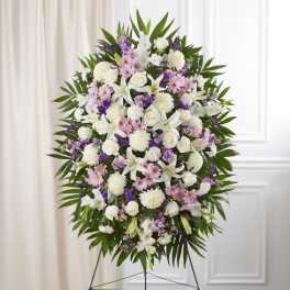 Tall standing spray of white lilies and roses with pink and purple flowers framed by green leaves on an easel.