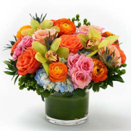 Low round arrangement of orange and pink roses, orchids, and ranunculus in a clear glass vase
