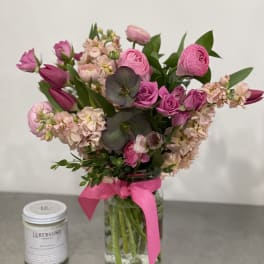 Pink flower arrangement with roses, tulips, and ranunculus in a glass jar with pink ribbon beside a white candle.