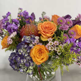Bouquet of orange roses, purple hydrangea, and mixed purple flowers in a glass vase