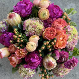 Vibrant round arrangement of fuchsia peonies, orange roses, peach ranunculus, hydrangeas, and protea.