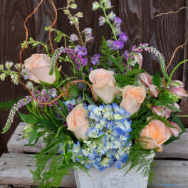 Mixed bouquet with pale roses, blue hydrangea, and purple blooms in a white vase