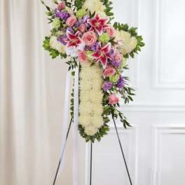 Standing floral cross with white base, pink roses, lilies, and lavender blooms on an easel