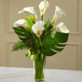 White calla lilies arranged in a clear glass vase with tropical leaves.