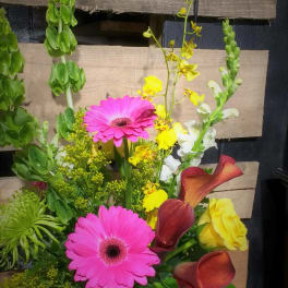Mixed bouquet with pink gerbera daisies, yellow blooms, and burgundy calla lilies in a glass vase.