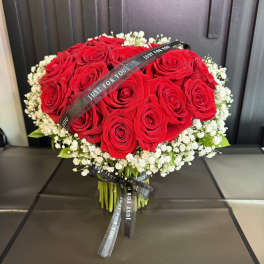 Hand-tied bouquet of red roses encircled by small white blooms with black Just For You ribbons.