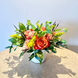 Low arrangement of orange and peach roses with yellow accents in a white ceramic vase on a round table