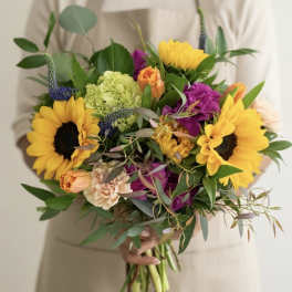 Hand-tied bouquet with bright yellow sunflowers, purple blooms, and mixed flowers held by a person in beige.
