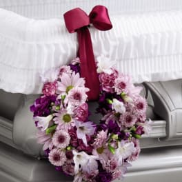 Pink and purple floral wreath with ribbon hanging from the corner of a white-lined casket