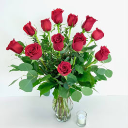 Red roses arranged in a clear glass vase with greenery and a small candle beside it
