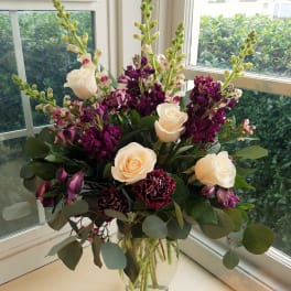 Tall arrangement of cream roses, purple flowers, and snapdragons in a clear glass vase