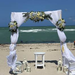 Beach wedding arch with white drapes and blue-and-cream floral arrangements