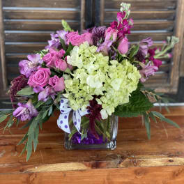 Pink roses and purple flowers in a square glass vase with a polka-dot ribbon.