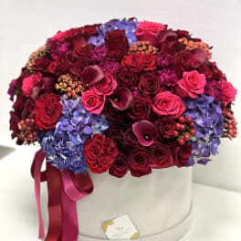 Round box arrangement of red roses, purple hydrangeas, and dark calla lilies