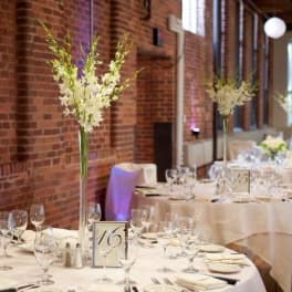 Tall white floral centerpieces on round banquet tables in a brick-walled room
