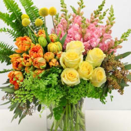 Bright arrangement of yellow roses, orange tulips, pink snapdragons, and ferns in a clear glass vase