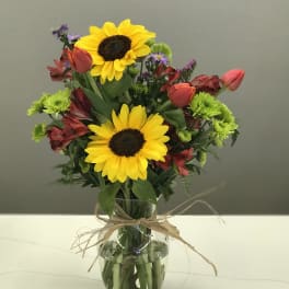 Sunflowers, tulips, and mixed blooms in a clear glass vase with raffia ribbon