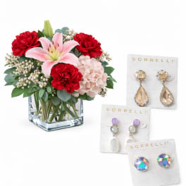 Bouquet of red and pink flowers in a square glass vase with three pairs of earrings beside it
