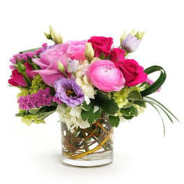 Low glass vase of pink roses, ranunculus, and mixed purple and white flowers in a compact arrangement