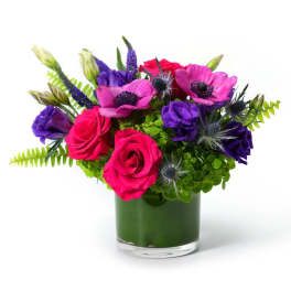 Low glass vase with hot pink roses, magenta anemones, purple flowers, and bright green blooms.