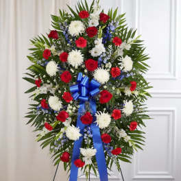 Patriotic standing spray with red roses, white mums, blue delphinium and a large blue ribbon on an easel.
