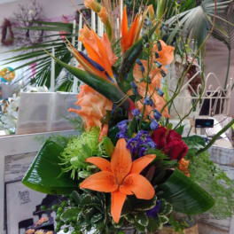 Tall tropical bouquet with orange lilies and purple accents in a glass vase