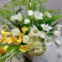 Tall arrangement of yellow calla lilies, white hydrangeas, tulips, and ranunculus in a white vase