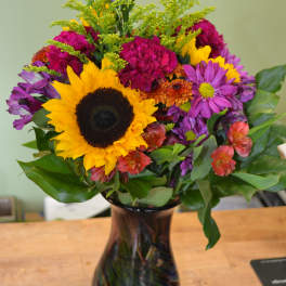 Mixed bouquet with a sunflower and purple, pink, and orange flowers in a dark glass vase