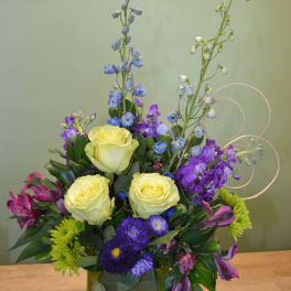 Mixed bouquet with yellow roses, purple flowers, and blue delphinium in a green vase