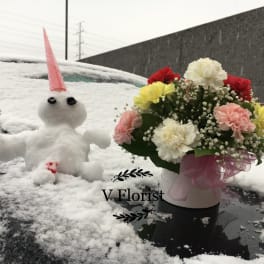 Round white box of mixed carnations with baby’s breath in snow beside a small snow figure.