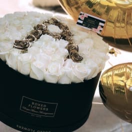 White roses arranged in a black round box with gold balloons nearby.