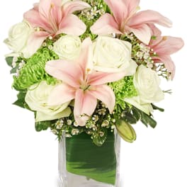 Compact arrangement of pink lilies, white roses, and green blooms in a clear rectangular vase.