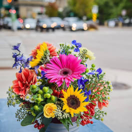 Bright mixed bouquet with sunflower, pink gerbera, and roses in a gray pot on a blue table outdoors.