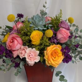 Mixed arrangement of pink and yellow roses, purple flowers, and a succulent in a red-brown vase