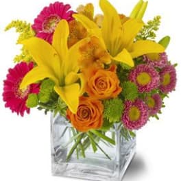 Bright mixed bouquet of yellow lilies, orange roses, and pink blooms in a clear glass cube vase