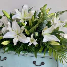 Low arrangement of white lilies with buds and abundant foliage on a pale blue dresser