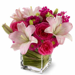 Low arrangement of pink lilies and roses in a clear glass cube vase with a leaf-wrapped base