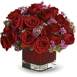 Red roses arranged in a square glass vase with small purple filler flowers.
