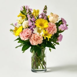 Mixed bouquet of yellow daisies, pink carnations, and purple flowers in a glass vase