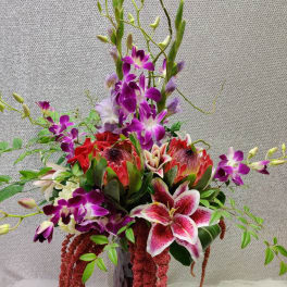 Tall tropical arrangement with purple orchids and pink lilies in a dark vase