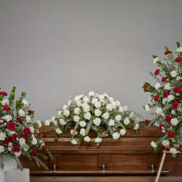 Red and white carnation sympathy set with casket spray and two standing arrangements beside a wooden casket