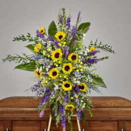 Standing spray of yellow sunflowers and purple flowers in front of a wooden casket