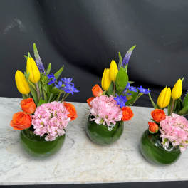 Three small floral arrangements in green glass vases with yellow, orange, pink, and blue flowers